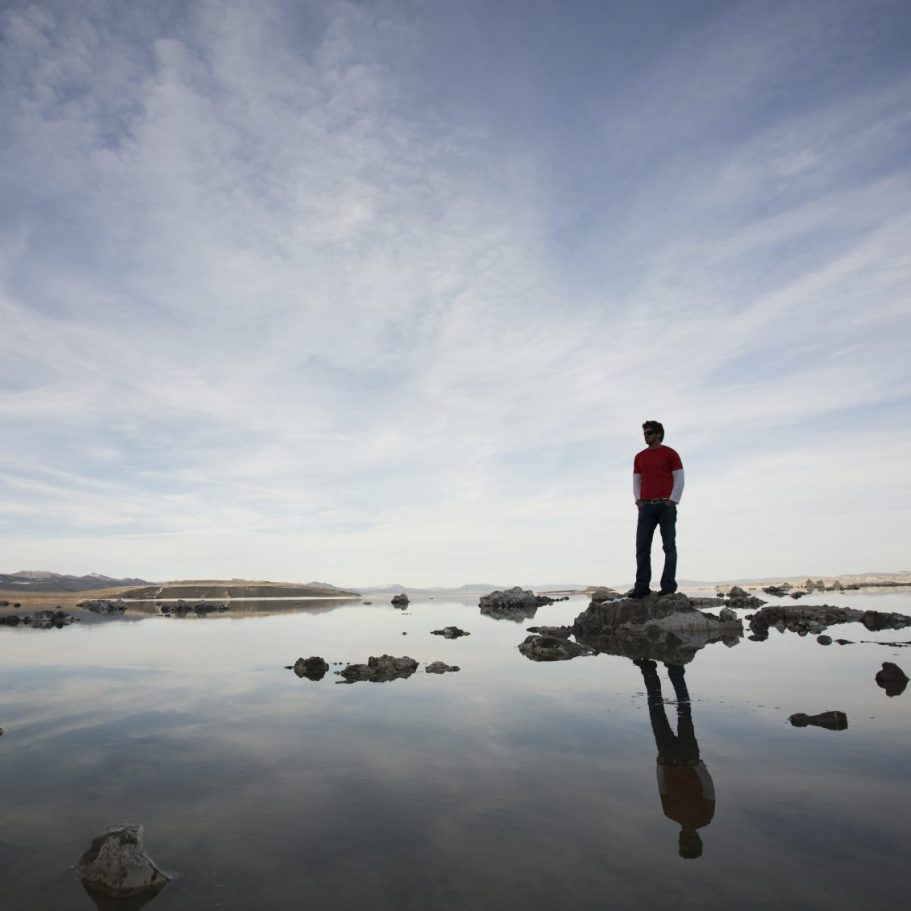 Junge steht auf einem Felsen am Wasser, reflektiert unter einem weiten Himmel.